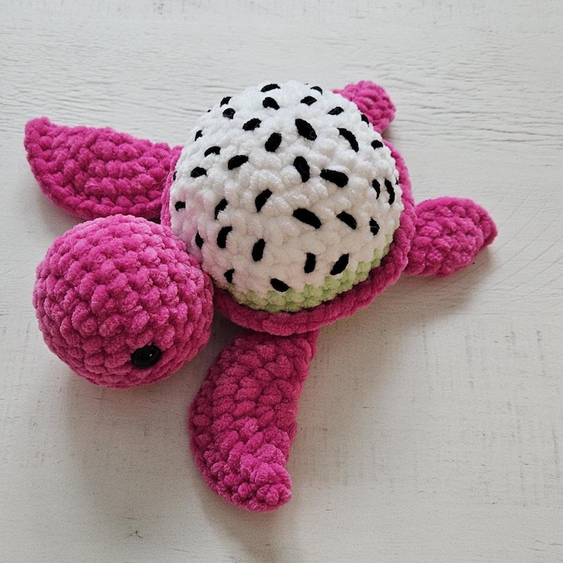 May include: A handmade crochet turtle plushie. The turtle's head is a white sphere with black spots, resembling a dragon fruit. The body, flippers, and tail are a vibrant pink. The plushie is made of soft, textured yarn.