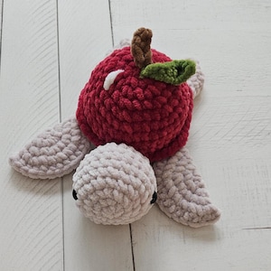 May include: A handmade crochet turtle toy with a red apple-shaped shell, brown stem, and green leaf. The turtle's head and flippers are light gray, with small black eyes. The toy is made of soft, textured yarn.
