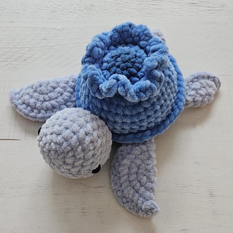May include: A handmade plush turtle in shades of blue and gray. The turtle features a textured, fluffy body, head, and flippers. The shell is a vibrant blue, and the head and flippers are a lighter gray. The turtle has small black eyes.