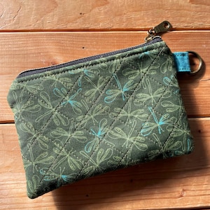May include: A rectangular quilted zipper pouch in olive green fabric featuring a repeating pattern of light green and teal dragonflies. The pouch includes a dark zipper closure and a small blue fabric tab with a metal D-ring attached to the side.