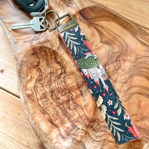 May include: A key fob with a colourful fabric design featuring a dark background with red, white, and green floral and architectural elements. The fob is attached to a car key and a house key with a metal ring.