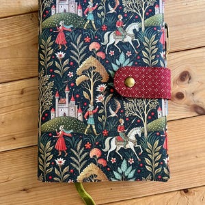 May include: A fabric-covered notebook featuring a dark blue background with a repeating pattern of castles, figures on horseback, and stylized trees. A red, polka-dot fabric strap with a brass snap closure secures the cover, and a green ribbon bookmark with a small metal key charm hangs from the bottom.