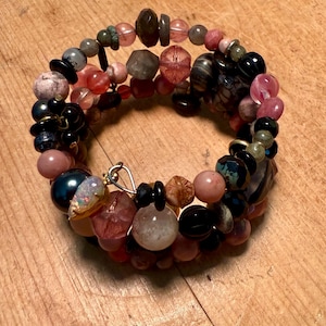 Puede incluir: Una pulsera de múltiples hilos con una variedad de cuentas coloridas en tonos rosa, negro, marrón y azul iridiscente. Las cuentas son de varias formas y tamaños, incluyendo redondas, facetadas e irregulares. Un cierre plateado asegura la pulsera.