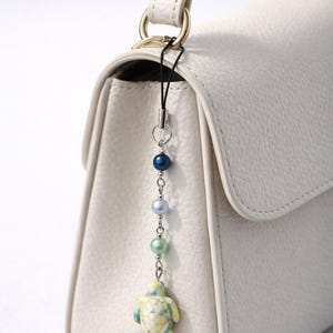 May include: A cream-colored handbag with a decorative charm. The charm features a small turtle pendant and a string of blue, green, and silver beads. The bag has a top handle and a gold-colored ring.