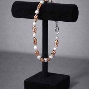 May include: A beaded bracelet with white and copper-coloured beads displayed on a black jewellery stand. The bracelet features alternating sections of white pearls and copper-coloured beads, connected by a silver clasp. The jewellery stand is black.
