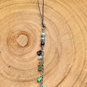 May include: A phone charm featuring a black cord loop attached to a silver-tone metal chain. The chain is adorned with three round beads in shades of light blue, medium blue, and dark blue, followed by five irregular stone chips and a turquoise turtle charm.