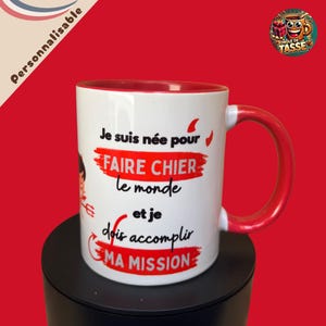 Peut inclure: Mug en céramique blanche avec intérieur et anse rouges. Le mug présente le texte français "Je suis née pour FAIRE CHIER le monde et je dois accomplir MA MISSION" en noir et rouge. Le mug est sur une surface noire sur fond rouge.