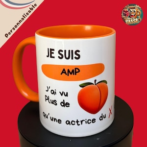 以下が含まれることがあります： オレンジ色の内側とハンドルが付いた白いセラミックマグ。マグには「JE SUIS AMP」と「J'ai vu Plus de qu'une actrice du X」のテキストと桃のグラフィックが描かれています。マグは赤い背景に対して黒い表面にあります。