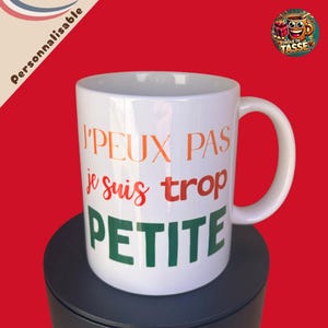 Peut inclure: Mug en céramique blanche avec une anse incurvée. Le mug affiche le texte "Je ne peux pas, je suis trop petite" en orange, rouge et vert. Le mug est posé sur une surface noire sur fond rouge.