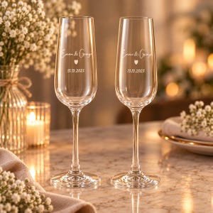 May include: Two clear glass champagne flutes featuring an etched design with the names Emma & George, a small heart icon, and the date 15.12.2025. The tall, slender glasses have a smooth, transparent finish and a stable circular base.