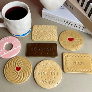 May include: A selection of ceramic biscuit-shaped coasters, featuring designs like custard cream, bourbon, and nice biscuits. A white mug with a blue stripe and a vase are also visible. Measurements are in cm.