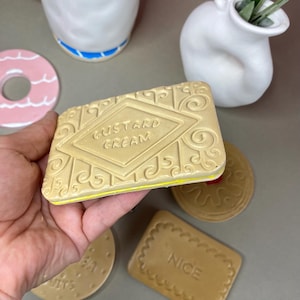 K&ouml;nnte beinhalten: Ein beigefarbenes Objekt in Form eines Custard Cream-Kekses mit dem gepr&auml;gten Schriftzug "Custard Cream" auf der Oberseite. Weitere keksf&ouml;rmige Objekte sind sichtbar, zusammen mit einer wei&szlig;en Tasse, einem rosa Donut und einer wei&szlig;en Vase.