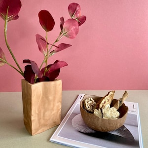 以下が含まれることがあります： バーガンディ色のユーカリの葉が入った紙袋のベースアレンジメント。茶色のボウルにはドライフラワーとスナックが入っており、雑誌の上に置かれています。背景は無地のピンク色です。