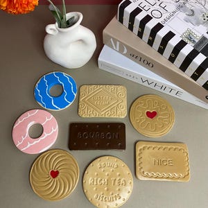 K&ouml;nnte beinhalten: Eine Sammlung von Untersetzern in Form von Keksen, darunter ein blauer Donut, ein rosa Donut und ein Custard Cream Keks. Weitere Untersetzer zeigen ein Herz, eine Spirale und Text. Eine wei&szlig;e Vase mit orangefarbenen Blumen und B&uuml;cher im Hintergrund.