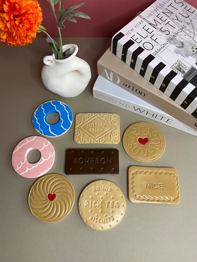 K&ouml;nnte beinhalten: Eine Sammlung von Untersetzern in Form von Keksen, darunter ein blauer Donut, ein rosa Donut und ein Custard Cream Keks. Weitere Untersetzer zeigen ein Herz, eine Spirale und Text. Eine wei&szlig;e Vase mit orangefarbenen Blumen und B&uuml;cher im Hintergrund.
