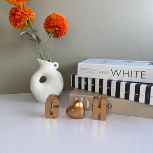 May include: A still-life arrangement featuring a white vase with a unique hole design, holding vibrant orange flowers. Gold-colored letters 'G' and 'H' and a heart-shaped object are displayed near stacked books.
