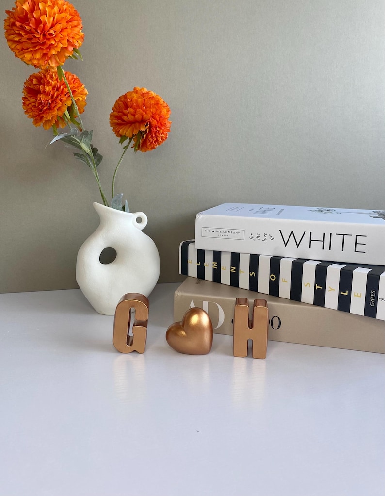 May include: A still-life arrangement featuring a white vase with a unique hole design, holding vibrant orange flowers. Gold-colored letters 'G' and 'H' and a heart-shaped object are displayed near stacked books.