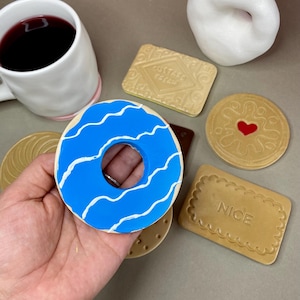 K&ouml;nnte beinhalten: Ein blauer Donut mit wei&szlig;en wellenf&ouml;rmigen Linien, umgeben von verschiedenen keksf&ouml;rmigen Gegenst&auml;nden. Eine wei&szlig;e Tasse mit rosa Rand und eine kleine wei&szlig;e Vase sind ebenfalls vorhanden. Die Gegenst&auml;nde sind auf einer neutralen Oberfl&auml;che angeordnet.