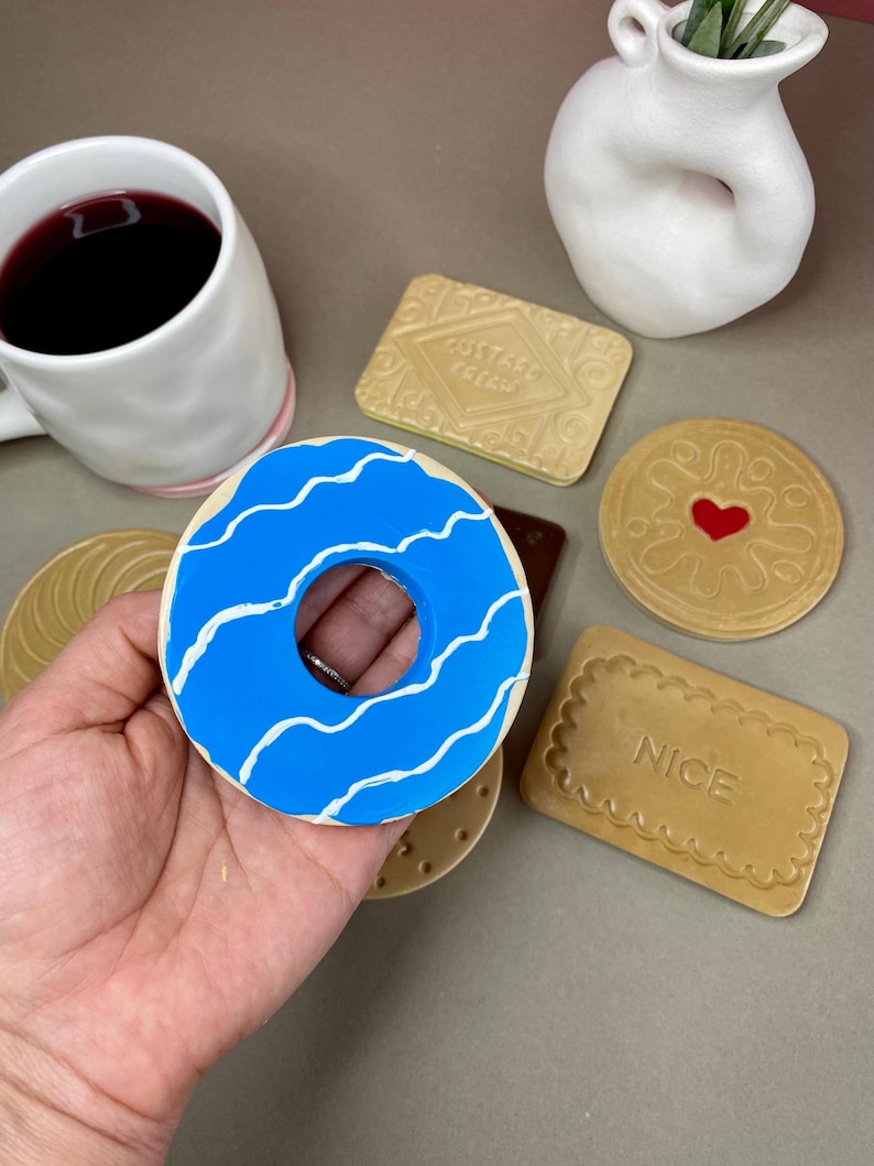 K&ouml;nnte beinhalten: Ein blauer Donut mit wei&szlig;en wellenf&ouml;rmigen Linien, umgeben von verschiedenen keksf&ouml;rmigen Gegenst&auml;nden. Eine wei&szlig;e Tasse mit rosa Rand und eine kleine wei&szlig;e Vase sind ebenfalls vorhanden. Die Gegenst&auml;nde sind auf einer neutralen Oberfl&auml;che angeordnet.