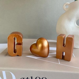 Puede incluir: Tres objetos decorativos de color cobre metálico: las letras "G" y "H", y una forma de corazón. Los objetos están colocados sobre un libro con el texto "at100". Un jarrón blanco está en el fondo.