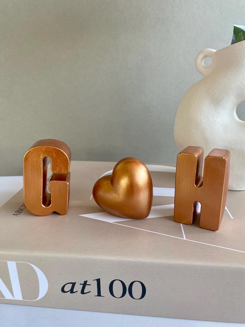 May include: Three metallic copper-colored decorative objects: the letters "G" and "H", and a heart shape. The objects are arranged on a book with the text "at100". A white vase is in the background.