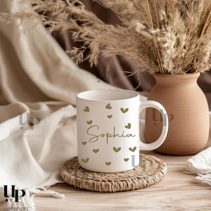 Peut inclure: Tasse en céramique blanche avec le nom "Sophia" en écriture cursive, ornée de cœurs dorés. La tasse est posée sur un dessous de verre tressé, avec un vase de plantes séchées en arrière-plan. La tasse est de taille standard.