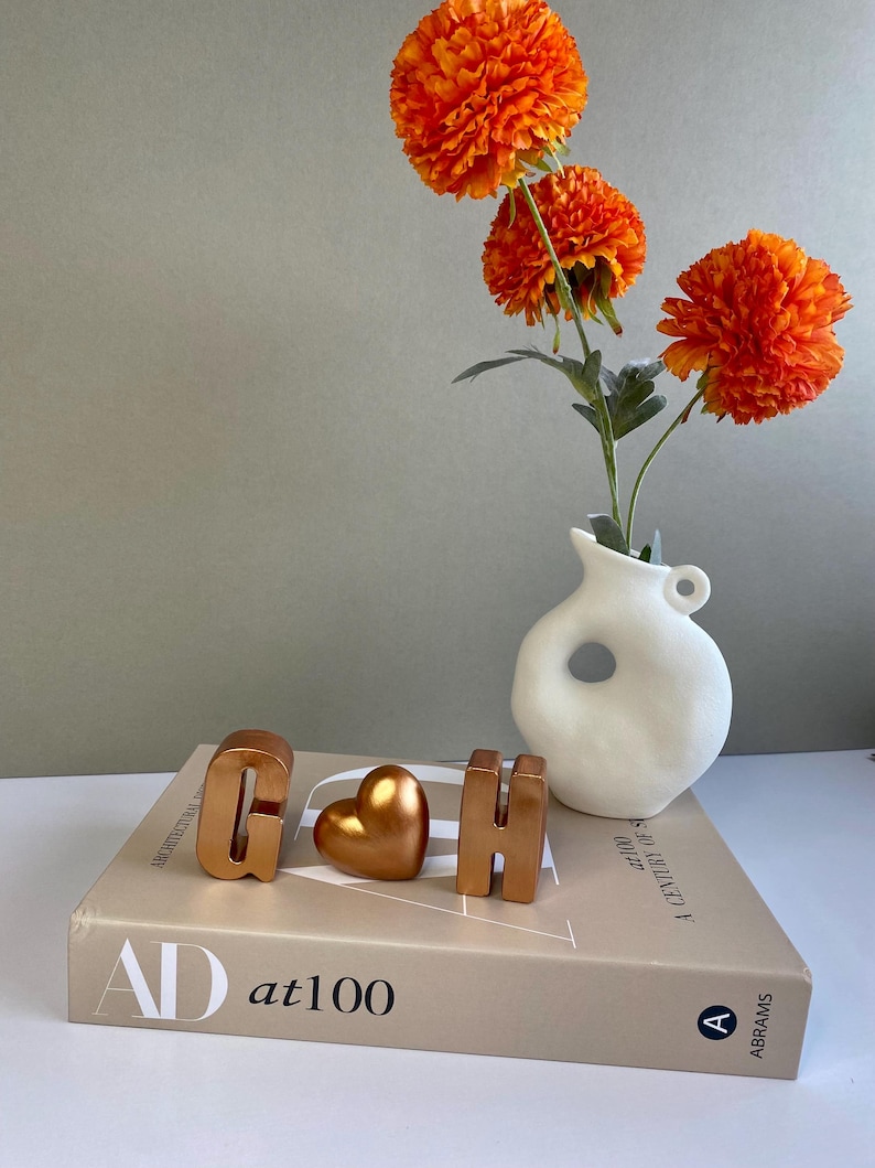 May include: A still life arrangement featuring a white vase with orange flowers, and gold-colored decorative letters and a heart on a book. The book has the text "AD at100" on the cover.
