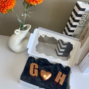 May include: A white box with a clear lid, containing gold-colored chocolate letters "G" and "H" and a heart-shaped chocolate. The box sits on black tissue paper. Orange flowers in a white vase are in the background.