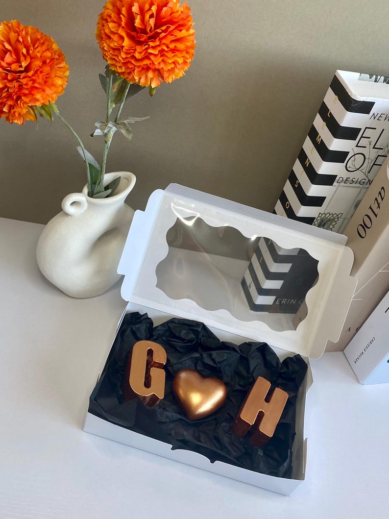 May include: A white box with a clear lid, containing gold-colored chocolate letters "G" and "H" and a heart-shaped chocolate. The box sits on black tissue paper. Orange flowers in a white vase are in the background.