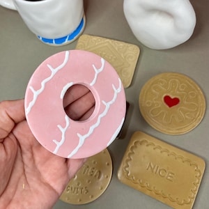 K&ouml;nnte beinhalten: Eine Sammlung von Untersetzern in Form von Keksen und einem Untersetzer in Donut-Form. Der Donut-Untersetzer ist rosa mit wei&szlig;en, geschwungenen Linien. Weitere Untersetzer zeigen ein Herz, ein Rechteck mit "NICE" und ein Rund mit "TEA CUITS".
