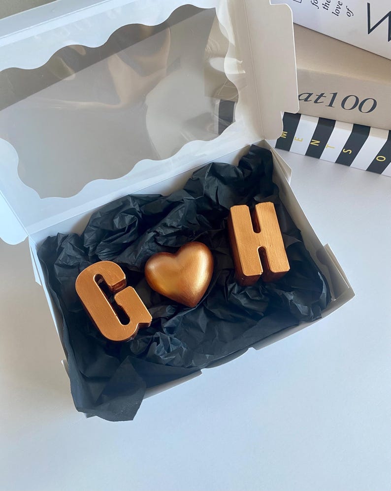 May include: A white gift box with a clear window contains a heart-shaped object and two letter-shaped objects. The objects are metallic copper-colored and rest on black tissue paper. The letters are "G" and "H".