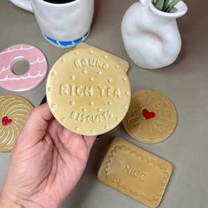 K&ouml;nnte beinhalten: Eine Sammlung von Untersetzern in Form von Keksen. Einer der Untersetzer tr&auml;gt die Aufschrift "ROUND RICH TEA BISCUITS". Weitere Designs sind ein rosa Donut, ein Herz und ein rechteckiger "NICE" Keks. Eine wei&szlig;e Tasse und Vase sind ebenfalls vorhanden.