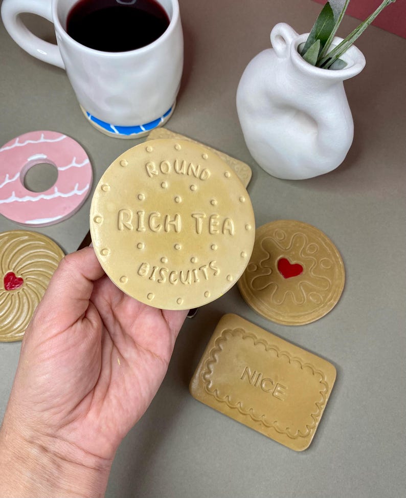 K&ouml;nnte beinhalten: Eine Sammlung von Untersetzern in Form von Keksen. Einer der Untersetzer tr&auml;gt die Aufschrift "ROUND RICH TEA BISCUITS". Weitere Designs sind ein rosa Donut, ein Herz und ein rechteckiger "NICE" Keks. Eine wei&szlig;e Tasse und Vase sind ebenfalls vorhanden.