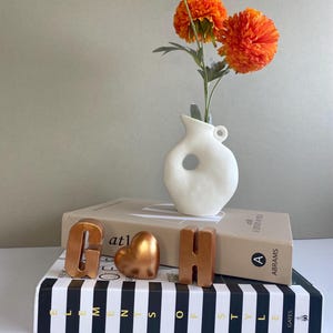 May include: A white ceramic vase with a circular cut-out holds three bright orange flowers. The vase sits atop two books, with copper-colored letters "G" and "H" and a heart-shaped ornament. The bottom book has black and white stripes.