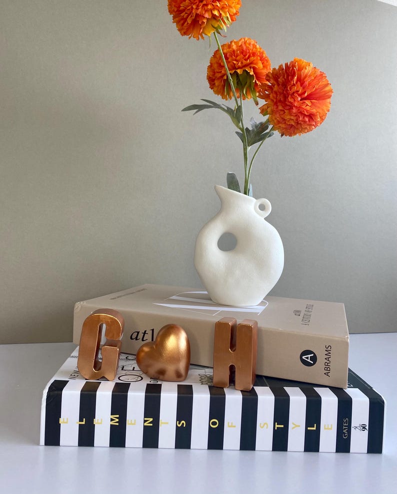 May include: A white ceramic vase with a circular cut-out holds three bright orange flowers. The vase sits atop two books, with copper-colored letters "G" and "H" and a heart-shaped ornament. The bottom book has black and white stripes.