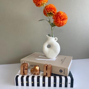 May include: A still-life arrangement featuring orange pom-pom flowers in a white vase with a unique shape. The vase sits atop a stack of books, with decorative copper-colored letters and a heart-shaped object. The books are on a black and white striped book.