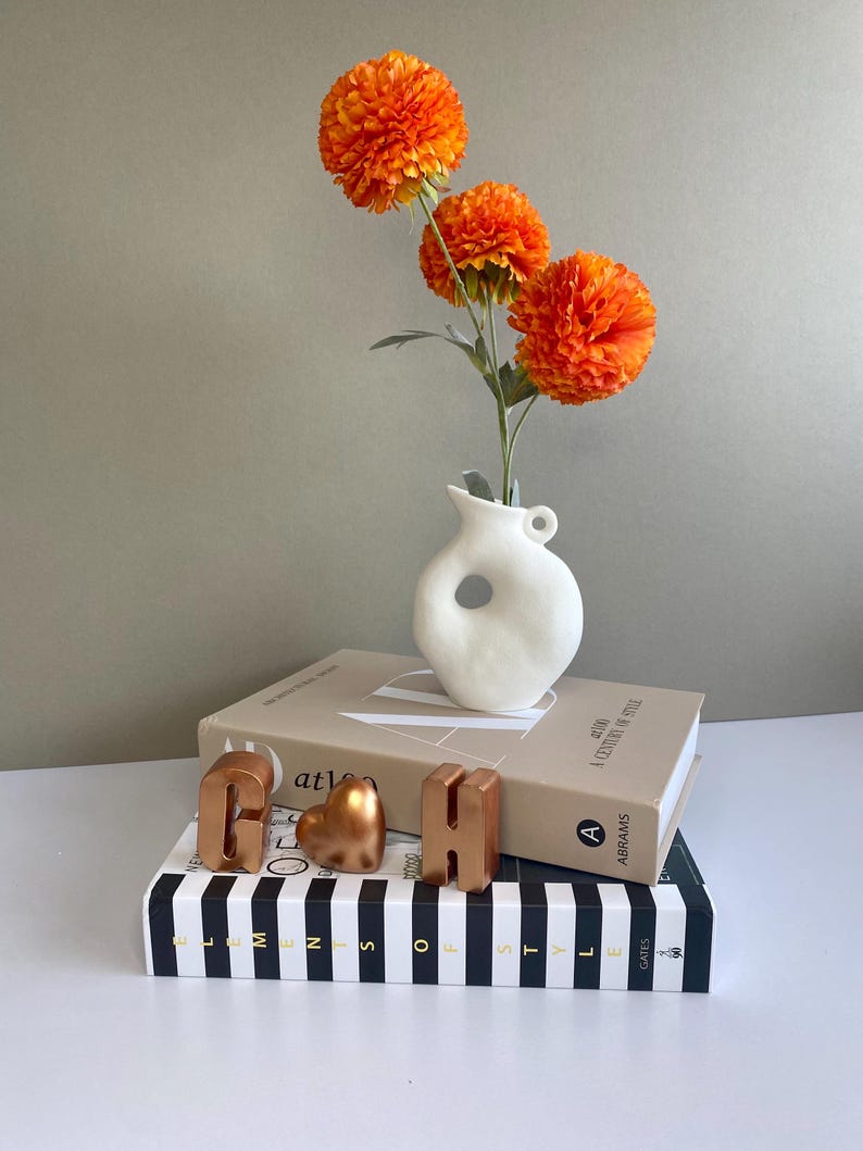 May include: A still-life arrangement featuring orange pom-pom flowers in a white vase with a unique shape. The vase sits atop a stack of books, with decorative copper-colored letters and a heart-shaped object. The books are on a black and white striped book.