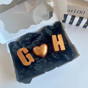May include: A white gift box with a clear window contains a heart-shaped object and two letter-shaped objects. The objects are metallic copper-colored and rest on black tissue paper. The letters are "G" and "H".