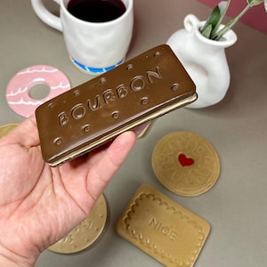 K&ouml;nnte beinhalten: Eine Hand h&auml;lt einen gro&szlig;en, braunen Keramik-Keks "Bourbon". Weitere Keramikkekse, ein rosa Donut, eine wei&szlig;e Tasse mit blauem Streifen und eine kleine wei&szlig;e Vase sind ebenfalls sichtbar. Die Tasse enth&auml;lt eine dunkle Fl&uuml;ssigkeit.