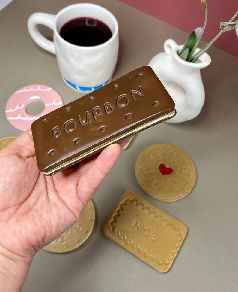 K&ouml;nnte beinhalten: Eine Hand h&auml;lt einen gro&szlig;en, braunen Keramik-Keks "Bourbon". Weitere Keramikkekse, ein rosa Donut, eine wei&szlig;e Tasse mit blauem Streifen und eine kleine wei&szlig;e Vase sind ebenfalls sichtbar. Die Tasse enth&auml;lt eine dunkle Fl&uuml;ssigkeit.