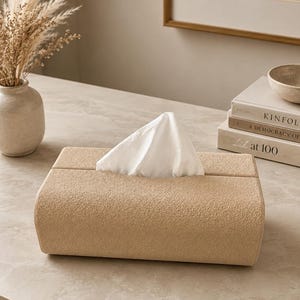 Op de afbeelding: Een rechthoekige tissueboxhouder met een getextureerde, beige, stofachtige afwerking. De houder heeft een centrale horizontale gleuf waaruit een enkele witte tissue naar boven wordt getrokken, waardoor een puntige vorm ontstaat.