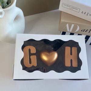 May include: A white box with a clear window displaying chocolate letters "G" and "H" flanking a gold heart. The chocolates are in a decorative arrangement. Books and a white vase are in the background.