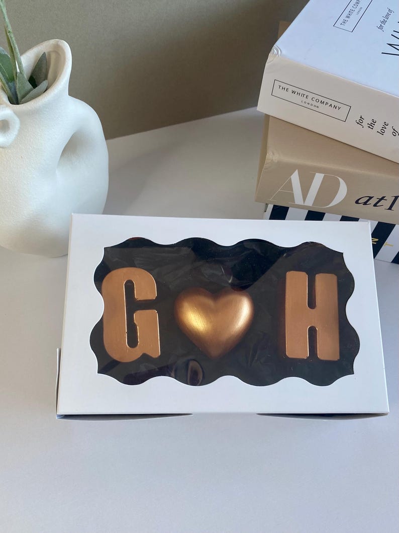May include: A white box with a clear window displaying chocolate letters "G" and "H" flanking a gold heart. The chocolates are in a decorative arrangement. Books and a white vase are in the background.
