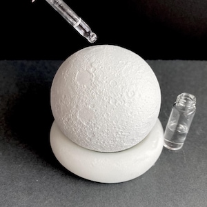 May include: A white, textured sphere, reminiscent of the moon, sits on a white, rounded base. A glass dropper dispenses liquid above the sphere. A small glass vial with liquid is also present. The background is dark.