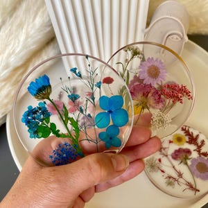 Puede incluir: Posavasos redondos de resina transparente con flores secas. Uno presenta hortensias azules y otros elementos florales azules y verdes. Otro tiene flores rosas, rojas y blancas. Un tercero tiene flores moradas y amarillas.