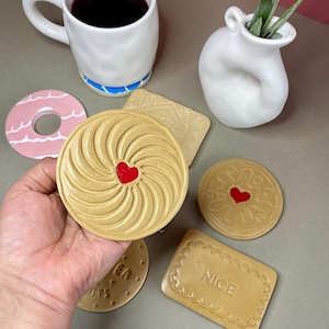 K&ouml;nnte beinhalten: Eine Sammlung von Keramikuntersetzern und einer Tasse. Die Untersetzer sind so gestaltet, dass sie wie Kekse und ein Donut aussehen, mit einer Vielzahl von Formen und Farben, darunter ein rosa Donut und beige Kekse mit roten Herzdetails. Eine wei&szlig;e Tasse mit blauem Streifen enth&auml;lt eine dunkle Fl&uuml;ssigkeit.