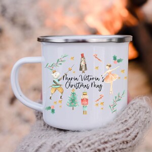 Puede incluir: Taza de esmalte blanco con borde y asa plateados, con un diseño festivo. La taza está decorada con ilustraciones de una bailarina, un cascanueces, un árbol de Navidad y el texto "Maria Victoria's Christmas Mug."