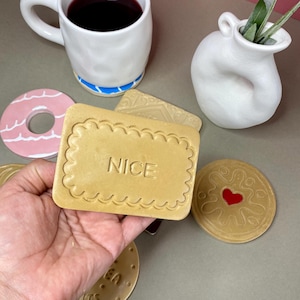 K&ouml;nnte beinhalten: Eine Sammlung von Keramikuntersetzern und einer Tasse. Die Untersetzer sind als Kekse gestaltet, einer davon mit dem Wort "NICE". Eine wei&szlig;e Tasse mit blauem Boden enth&auml;lt eine dunkle Fl&uuml;ssigkeit.