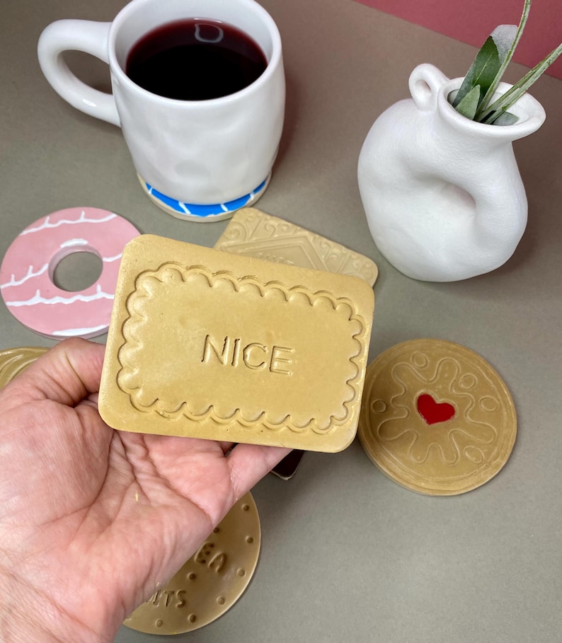 K&ouml;nnte beinhalten: Eine Sammlung von Keramikuntersetzern und einer Tasse. Die Untersetzer sind als Kekse gestaltet, einer davon mit dem Wort "NICE". Eine wei&szlig;e Tasse mit blauem Boden enth&auml;lt eine dunkle Fl&uuml;ssigkeit.