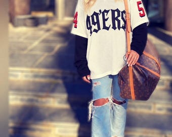 May include: A white mesh V-neck jersey with black 49ERS text and red number 45 accents, worn over a black long-sleeve shirt. Paired with light blue distressed denim jeans featuring large knee rips and tan suede lace-up boots, plus a brown monogram duffel bag.